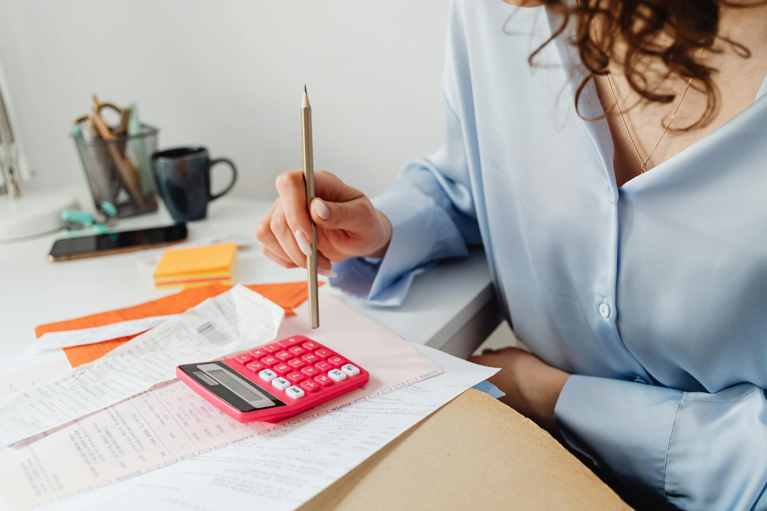A woman is calculating expenses and bookkeeping for her business by using a calculator and papers at her desk.