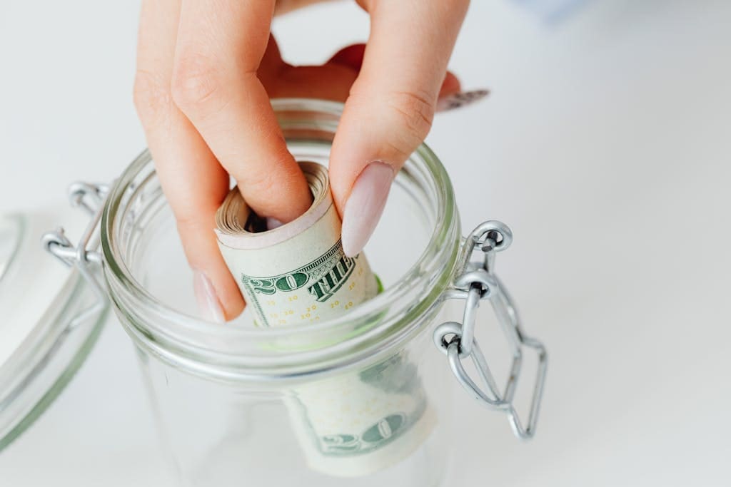 A close-up of a hand placing rolled dollars into a glass jar, symbolizing an emergency fund