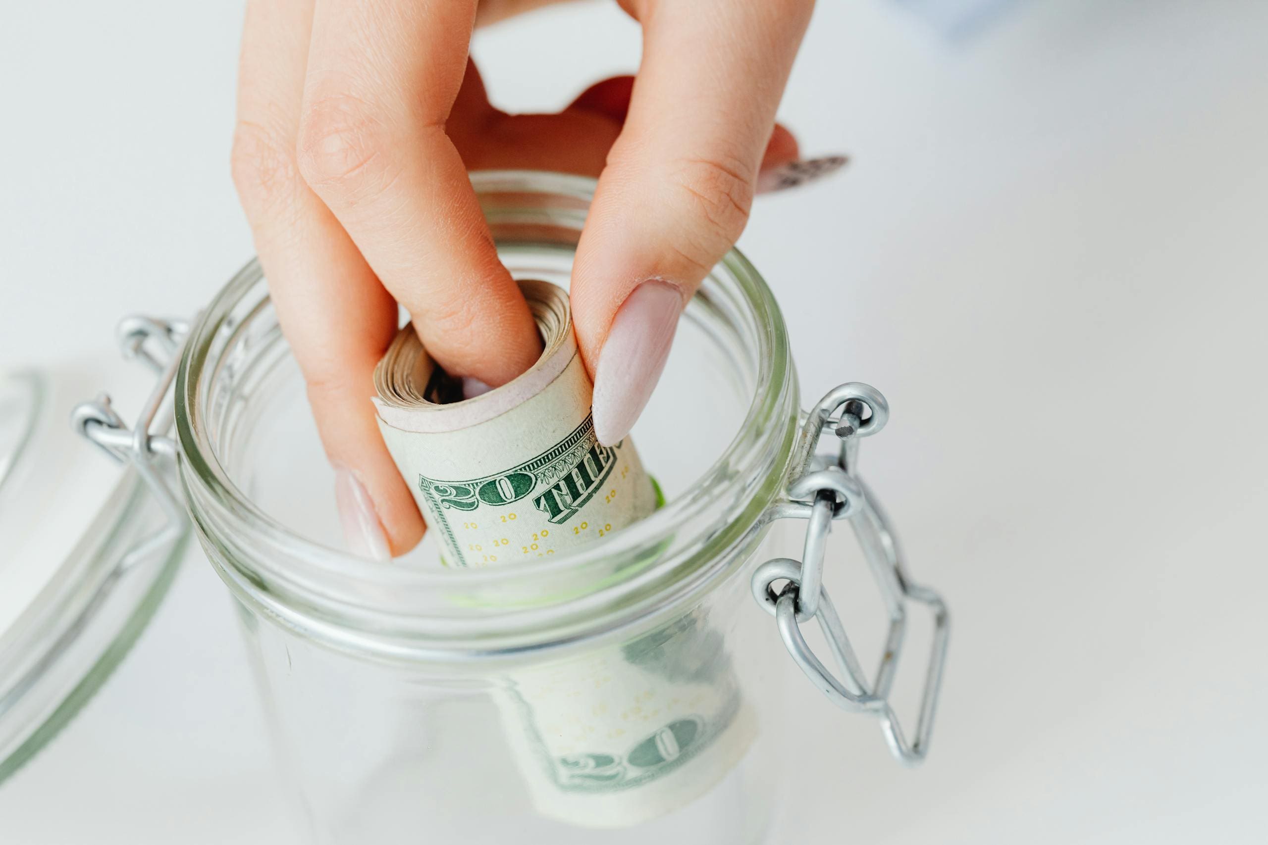 A close-up of a hand placing rolled dollars into a glass jar, symbolizing an emergency fund