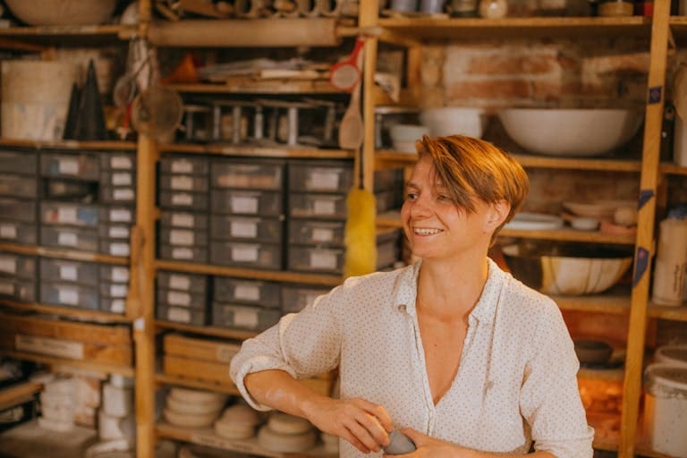 A smiling artisan in a pottery workshop surrounded by tools and shelves, creating ceramic art after scheduling a call with Two Rivers Bookkeeping.