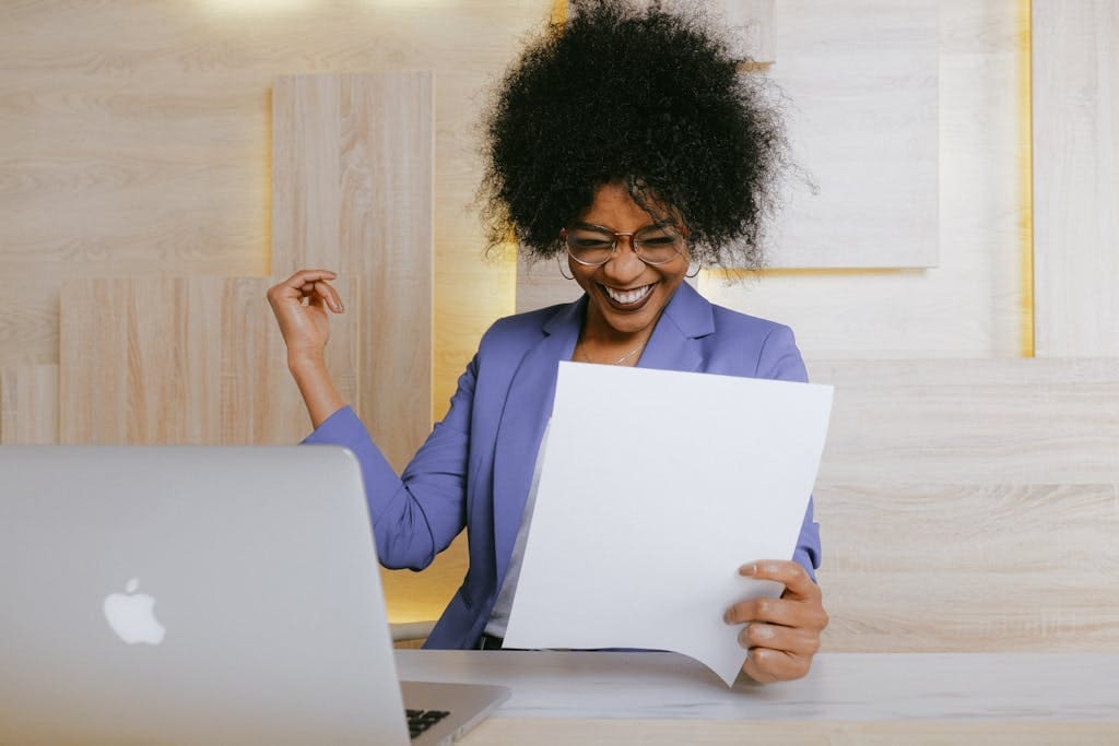 A cheerful woman celebrates her online bill paying success at work, looking at a document in an office setting.